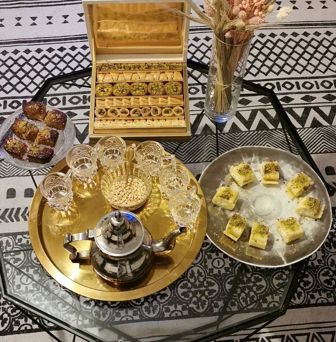 Table dressée vue de dessus : coffret de baklawa ouvert, plateau doré avec théière, verres et pignons de pin, assiette de halawet el jibn à la pistache et pâtisseries